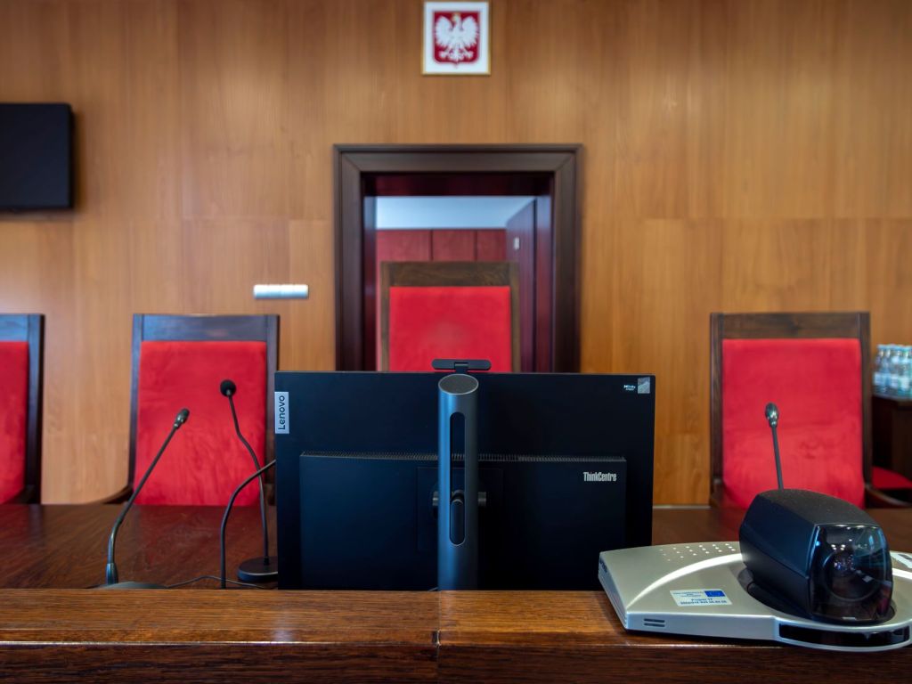 Vista del interior de un tribunal con una computadora y micrófonos en el mostrador principal, sillas rojas al fondo.